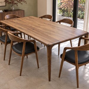 Three-quarter view showing the rounded walnut table in an olive-toned garden dining room. Styled with walnut chairs and black seats. This listing includes the dining table only; chairs and decorative accessories are not included.