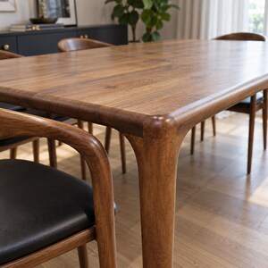 Close view of the rounded tabletop corner, leg shoulder and nearby walnut armchair. Styled with walnut chairs and black seats. This listing includes the dining table only; chairs and decorative accessories are not included.