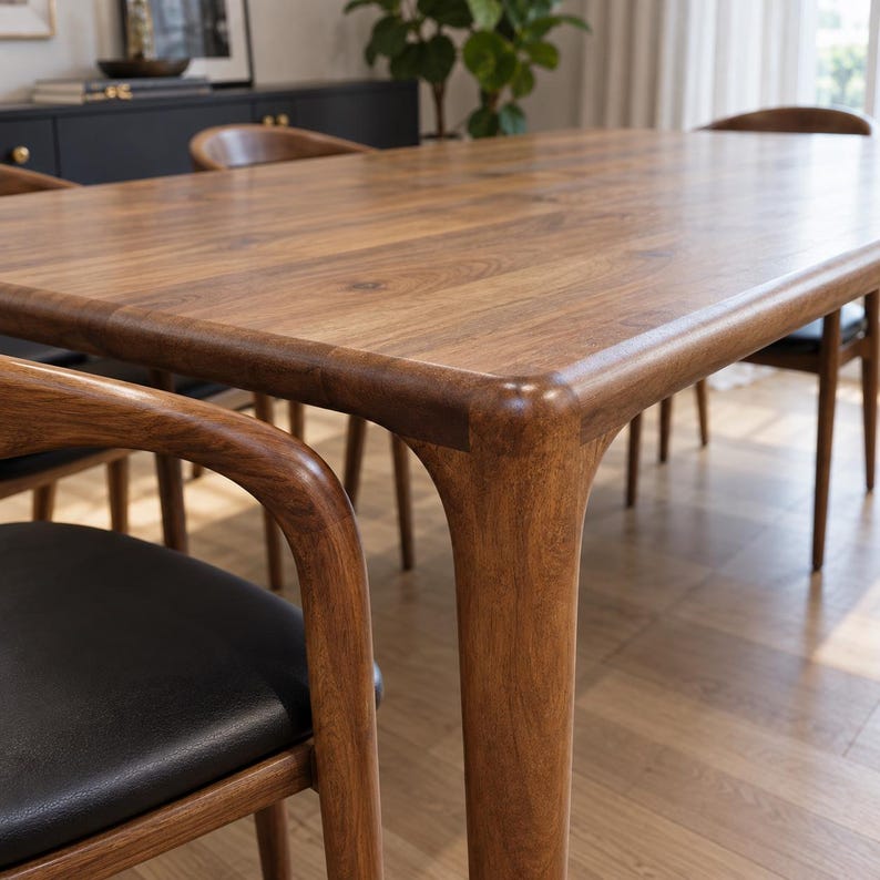 Close view of the rounded tabletop corner, leg shoulder and nearby walnut armchair. Styled with walnut chairs and black seats. This listing includes the dining table only; chairs and decorative accessories are not included.