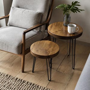 May include: Two round side tables with natural wood grain tops and black metal hairpin legs. The taller table holds a clear glass vase with green leaves and a white ceramic mug. The tables are positioned next to a light grey upholstered chair.