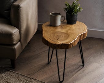 May include: A side table featuring a natural live-edge wood slab top with visible grain patterns, supported by three thin black metal hairpin legs. A light-coloured ceramic mug and a small green potted plant sit on the wooden surface.