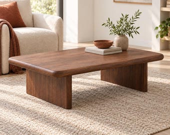 May include: A low-profile rectangular coffee table crafted from dark brown wood with rounded edges and two solid slab legs. The table surface holds a stack of two light-colored books, a small terracotta-colored bowl, and a ceramic vase with greenery.