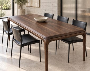 May include: A rectangular dining table made of dark brown wood with rounded corners and four tapered legs, accompanied by six chairs featuring black leather upholstery and thin black metal frames, with a speckled bowl centered on the table.