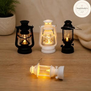 May include: Four miniature hurricane lanterns with LED lights. Three lanterns stand upright in black, white, and black, while one white lantern lies horizontally. Each features a clear glass centre, a metal handle, and a warm glowing light source.