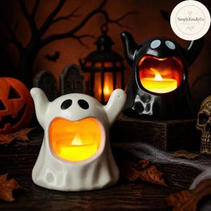 Puede incluir: Dos portavelas de cerámica con forma de fantasma, uno blanco y otro negro, cada uno con una boca abierta que contiene una vela LED amarilla brillante. Los fantasmas tienen los brazos levantados y ojos ovalados negros para Halloween.