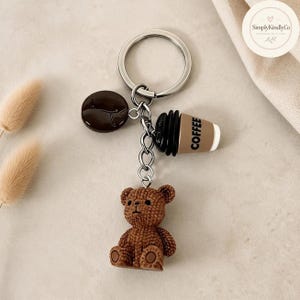 Pode incluir: Um porta-chaves de metal prateado com três pendentes: um grão de café castanho escuro, um copo de café bege e preto com a palavra COFFEE impressa na lateral e uma figura de urso de peluche castanho texturado, presos a um anel metálico.