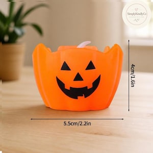 Puede incluir: Un recipiente de plástico naranja con forma de calabaza de Halloween y un diseño de ojos y boca triangulares negros. El recipiente mide 4 cm de alto y 5,5 cm de ancho. Una pequeña lengüeta de color blanco rosáceo sobresale de la parte superior.