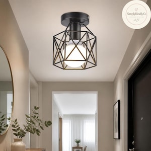 Puede incluir: Una lámpara de techo semiempotrada de metal negro con un diseño de jaula geométrica. El accesorio tiene un casquillo central cilíndrico que sostiene una bombilla blanca redonda, creando un aspecto estructurado para la iluminación interior.