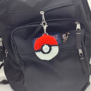 May include: A black backpack with a red and white crocheted Pokeball keychain attached to the zipper. The keychain is hanging from a silver metal loop.