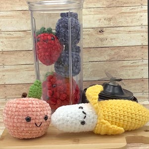 May include: A set of crocheted plush fruits and berries, including a peach, banana, and strawberries, in a clear blender container. The fruits have cute faces and are made with soft, fuzzy yarn.