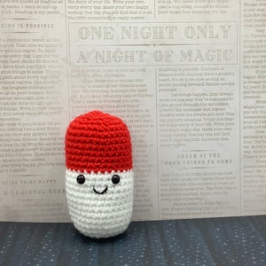 May include: A crocheted pill-shaped plushie with a red top and white bottom. The plushie has a smiling face with black eyes and a mouth. The plushie is sitting on a piece of paper with text that reads "One Night Only A Night of Magic" and "Dream of the Good Things to Come".