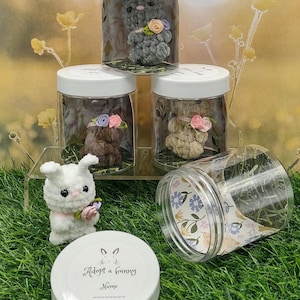 May include: Three clear glass jars with white lids, each containing a crocheted bunny with a flower. A white jar with a lid that says "Adopt a bunny" and a space for a name is in the foreground. The jars are on a green surface.