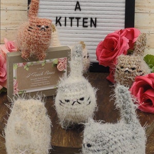 May include: Five crocheted plush kittens in various colors, including gray, white, and brown, with fluffy fur. The kittens are arranged on a wooden surface with a sign that reads "Adopt a Kitten".