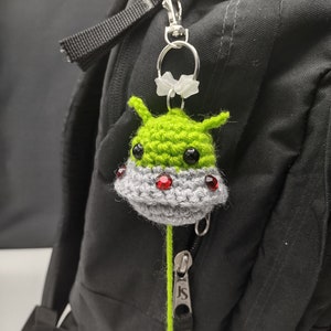 May include: A green crocheted alien with red eyes and a gray crocheted spaceship attached to a green zipper pull. The zipper pull is attached to a black backpack.