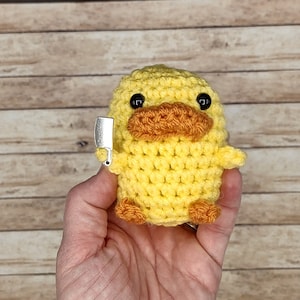 May include: A crocheted yellow duck holding a small silver knife. The duck has black eyes and an orange beak.