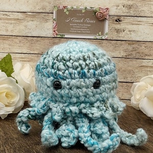 May include: A crocheted blue and green octopus with black eyes. The octopus is sitting on a wooden surface.