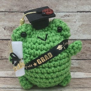 May include: Green crocheted frog wearing a black graduation cap with a red ladybug on top. The frog is holding a white scroll with a gold 2023 charm and a black ribbon. The frog has a black ribbon around its body that says "Congrats Grad" in gold letters.