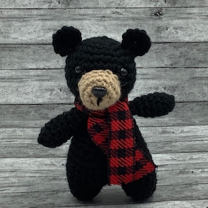 May include: A black crocheted bear wearing a red and black plaid scarf. The bear has a brown nose and black eyes.