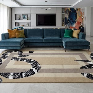 May include: A rectangular area rug featuring a beige and grey geometric border design with a large, winding black and white snake motif that curves across the surface. The rug is positioned on a light-coloured floor in front of a large blue sofa.