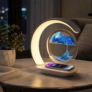 May include: A white crescent-shaped desk lamp with an integrated circular sand art display featuring blue and white sand. The base of the lamp functions as a wireless charging pad, currently holding a smartphone with a visible battery icon.