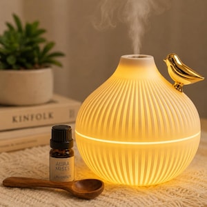 Puede incluir: Un difusor de aceites esenciales esférico de color blanco con texturas estriadas verticales, que emite una fina niebla. Una pequeña figura de pájaro de color dorado pulido está unida al lateral. Junto a él hay un frasco de vidrio marrón con la etiqueta Aura Mists Lavender.