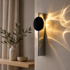 Peut inclure: Une applique murale composée d'une base rectangulaire métallique verticale et d'un disque noir circulaire. La lampe émet une lueur dorée et chaude qui projette des motifs lumineux fluides et distincts sur la surface du mur adjacent.