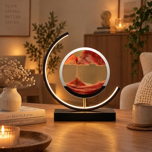 May include: A circular sand art lamp featuring a black base and a curved frame with integrated LED lighting. The central glass disc contains layers of red and white sand, creating a mountain-like landscape as the sand settles inside the frame.