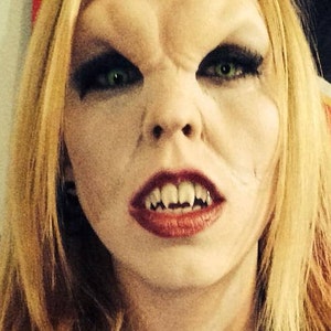 Könnte beinhalten: Nahaufnahme eines Gesichts mit Vampir-Make-up. Das Gesicht ist blass mit übertriebenen Merkmalen, darunter spitze Eckzähne, dunkles Augen-Make-up und roter Lippenstift. Blondes Haar umrahmt das Gesicht, und die Person blickt direkt in die Kamera.