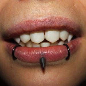 May include: Close-up of a person's mouth with a black lip ring and a black spike piercing. The person has a small, pointed tooth on the bottom left side of their mouth.