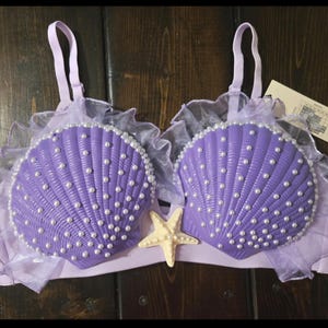 May include: A lavender bralette featuring two molded purple seashell cups adorned with white faux pearls. The garment includes light purple ruffled tulle trim along the top edges, adjustable shoulder straps, and a cream-colored starfish accent.