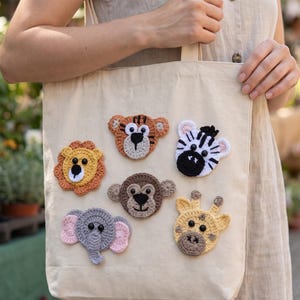 Puede incluir: Un bolso de tela beige con seis aplicaciones de animales tejidas a ganchillo en la parte frontal. Los diseños incluyen un león, un tigre, una cebra, un mono, un elefante y una jirafa, elaborados con hilo en tonos amarillo, naranja, negro, blanco y gris.