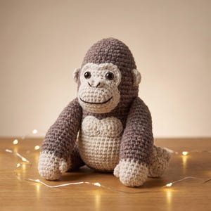 May include: A handmade crochet gorilla stuffed animal featuring dark brown yarn for the head, arms, and legs, with light beige yarn for the face, chest, and hands. The gorilla has two black plastic eyes and a stitched black mouth line.