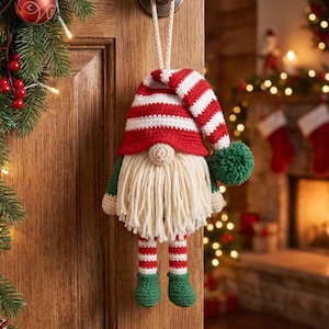 May include: A crocheted gnome ornament featuring a red and white striped hat with a green pom-pom, a beige nose, a long cream-colored yarn beard, and red and white striped legs ending in green crocheted boots, suspended by a white rope loop.