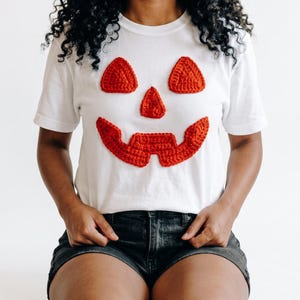 May include: A white short-sleeved t-shirt featuring a large, orange, crocheted jack-o'-lantern face applique on the front. The design consists of two triangular eyes, a triangular nose, and a wide, jagged, toothy grin made of textured yarn.