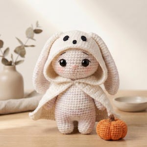 Könnte beinhalten: Eine cremefarbene Häkelpuppe mit Kapuzenumhang, zwei schwarzen Punkten auf der Stirn, großen dunklen Augen und rosigen Wangen. Die Puppe wird von einem kleinen, strukturierten orangefarbenen Häkelkürbis begleitet, der zu ihren Füßen liegt.
