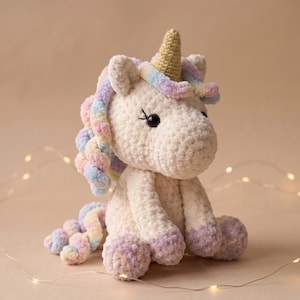 Puede incluir: Un peluche de unicornio tejido a ganchillo en color crema con cuerpo de hilo texturizado, un cuerno dorado y una melena y cola en tonos pastel multicolor. El unicornio tiene ojos negros de seguridad con pestañas bordadas y cascos morados.