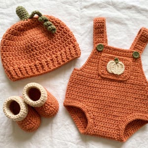 Op de afbeelding: Een handgehaakte set bestaande uit een oranje pompoenvormige muts met een groene steel, een paar oranje slofjes met crèmekleurige boorden en een oranje romper met twee groene knopen en een kleine crèmekleurige pompoenapplicatie.