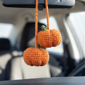 May include: Two small, orange, crocheted pumpkins with green stems hanging from a matching orange yarn cord. The pumpkins are suspended from a car rearview mirror, featuring a textured, hand-stitched yarn construction with visible ridges.