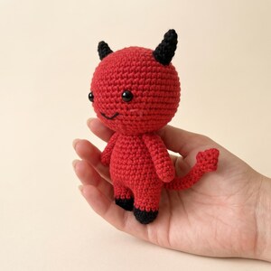 May include: A small, handmade crochet doll shaped like a red devil. The figure features a round head with two black horns, black bead eyes, and a stitched smile. It has a red body, black feet, and a long, pointed red tail with a spade-shaped tip.