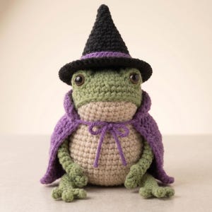 May include: A crocheted frog figurine featuring a green head and limbs with a light tan belly. The frog wears a pointed black hat with a purple band and a purple cape tied at the front with a matching purple string. The frog has dark eyes.