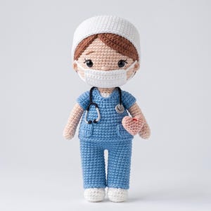 Op de afbeelding: Een gehaakte pop verkleed als zorgverlener, met een witte muts, een wit mondkapje en een blauw scrub-setje. De pop heeft bruin haar, een zwarte stethoscoop om de nek en houdt een klein, lichtroze gehaakt hartje vast in de hand.