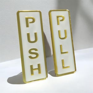 Puede incluir: Dos letreros rectangulares para puerta con fondo blanco y borde dorado. Un letrero presenta la palabra PUSH en letras doradas, y el otro letrero presenta la palabra PULL en letras doradas. Ambos letreros tienen un acabado liso y reflectante.