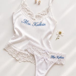 May include: A white camisole and matching thong set featuring delicate lace trim and the embroidered blue text Mrs. Kathrin. The camisole has thin straps and a V-neckline, while the thong includes small bows and lace detailing.