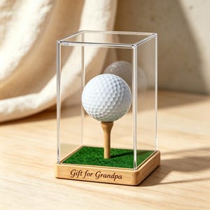 May include: A white golf ball sits on a wooden tee inside a clear acrylic display case. The base of the case is made of light wood and features the engraved text "Gift for Grandpa" on the front, with a green artificial grass surface inside.