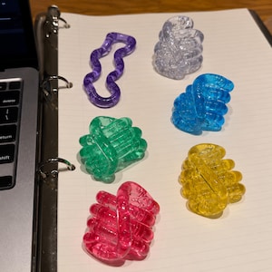 May include: A collection of six translucent plastic fidget toys in various colors including purple, clear, blue, green, yellow, and pink. Each toy consists of a series of connected, articulated segments that can be manipulated into different shapes.