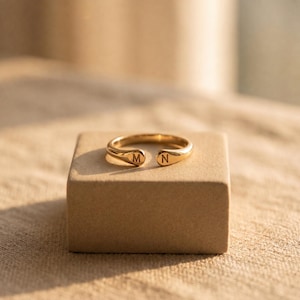 May include: A gold-toned open ring featuring two rounded ends, each engraved with a single capital letter. One end displays the letter M, while the other end displays the letter N. The ring has a smooth, polished finish and a simple, continuous band.