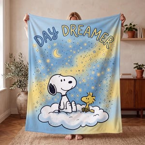 May include: A soft fleece blanket featuring a light blue and yellow gradient background with a starry night sky, a crescent moon, and the text DAY DREAMER. Snoopy and Woodstock are sitting together on a large white cloud in the center.