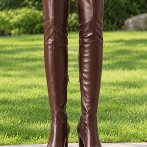 May include: A pair of over-the-knee boots in a smooth dark brown finish. These boots feature a pointed toe, a high block heel, and a vertical side zipper closure that runs along the inner shaft. The material appears flexible and form-fitting.