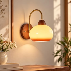 May include: A wall-mounted lamp featuring a circular dark wood base, a curved brass arm, and a rounded glass shade with a warm white to amber gradient. The light fixture emits a soft glow, with a dark wood accent piece connecting the shade to the arm.