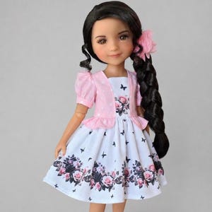 Könnte beinhalten: Eine Puppe mit langem, dunkelbraunem Haar in einem seitlichen Zopf, bekleidet mit einem rosa gepunkteten Bolero über einem weißen Kleid. Das Kleid zeigt ein Blumenmuster aus rosa und schwarzen Rosen am Saum, schwarze Schmetterlinge und eine rosa Blume.
